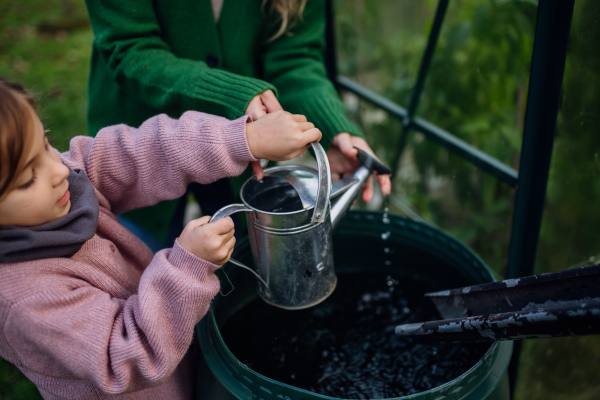 Girl and mother scooping rainwater from barell under downspout, going to water plants in garden. Concept of water conservation in garden.