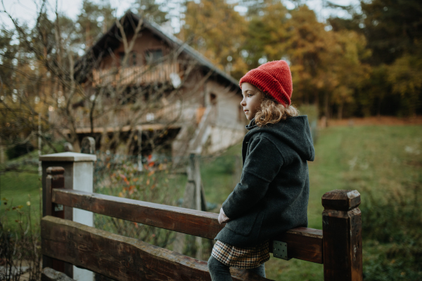 Young girl in warm clothes sitting on aged wooden fence or wooden gate, looking at autumn nature.