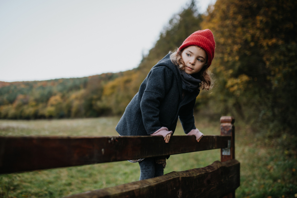 Young girl in warm clothes sitting on aged wooden fence or wooden gate, looking at autumn nature.