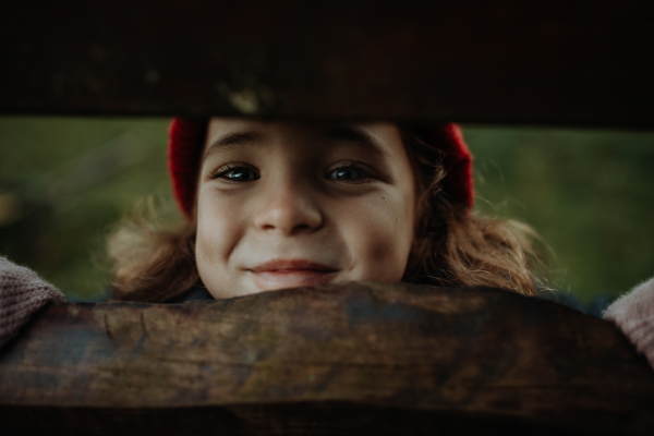 Portrait of cute little girl hiding behind wooden fence, looking between wooden boards.