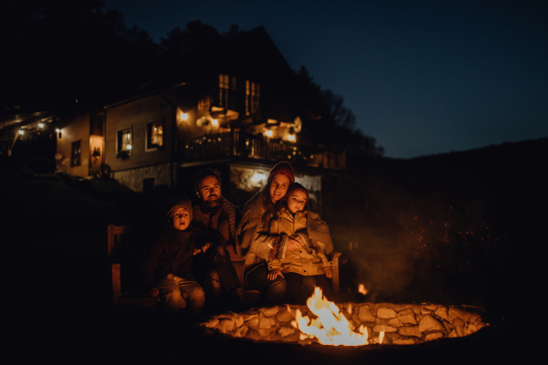 The family is warming up by an outdoor fire pit, sitting in the evening warmth of the fire, spending quality family time together.