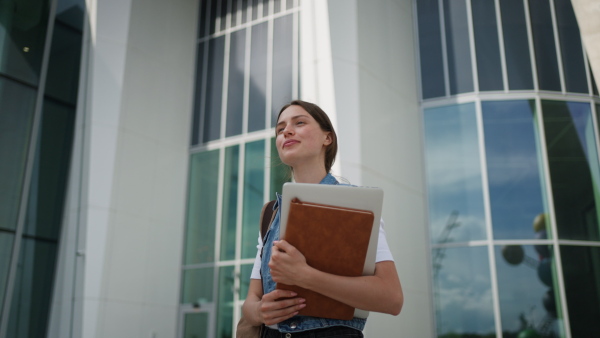 A female university student in front of university building or campus, backpack on back. Holding textbook, heading to exam, or lecture, study group.