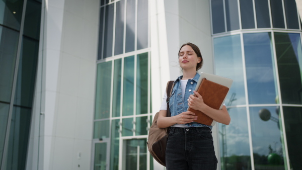 A female university student in front of university building or campus, backpack on back. Holding textbook, heading to exam, or lecture, study group.