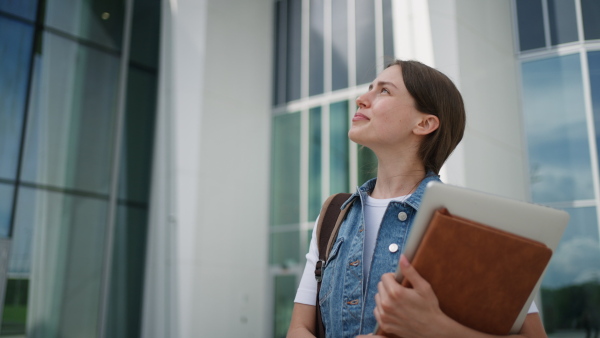 A female university student in front of university building or campus, backpack on back. Holding textbook, heading to exam, or lecture, study group.