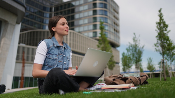 A female student sitting on grass in front of university building or campus. Working on laptop. Studying outdoors.