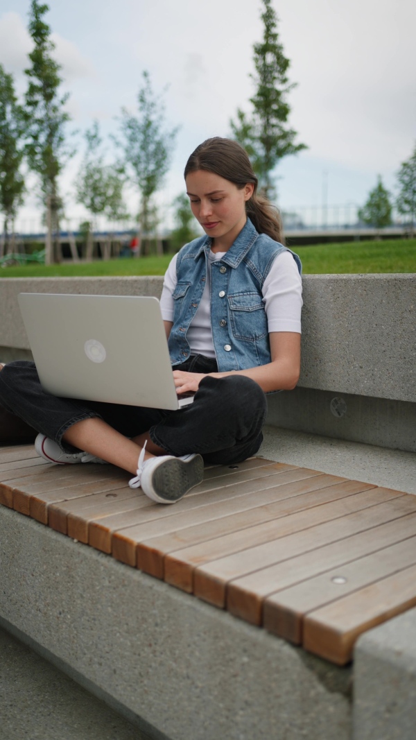 A female student sitting on bench in front of university building or campus. Working on laptop. Studying outdoors.