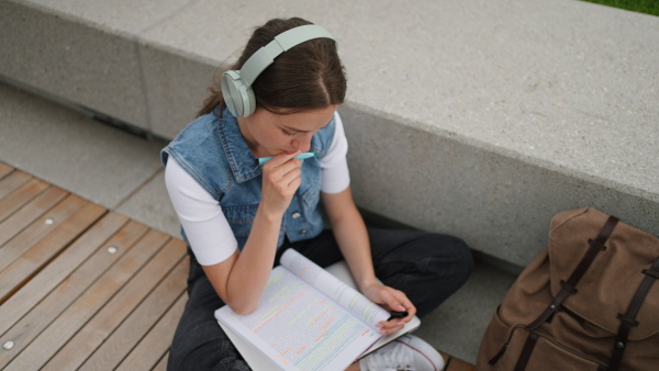 An university student with headphones sitting on ground in park, at campus and reading textbook. Studying before exam.