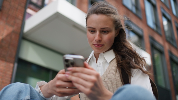 An university student sitting in front of university building. Scrolling her smarthpone.