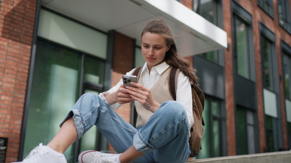 An university student sitting in front of university building. Scrolling her smarthpone.