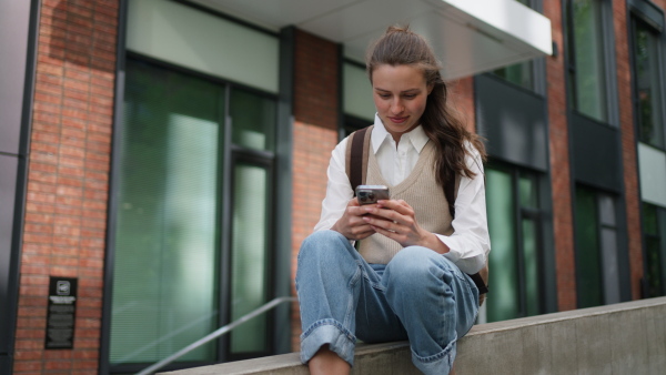 An university student sitting in front of university building. Scrolling her smarthpone.