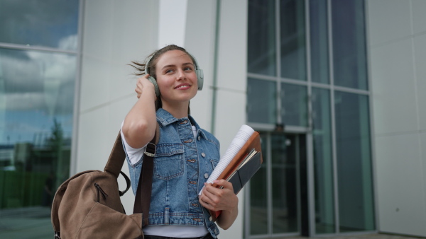 A female university student with headphones in front of university building or campus, backpack on back. Holding textbook, heading to exam, or lecture, study group.