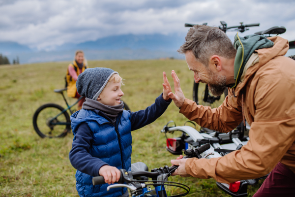 Young family with little children preparing for bicycle ride in nature, putting off bicycles from car racks. Father and son high five, looking forward to ride together. Concept of healthy lifestyle and moving activity.