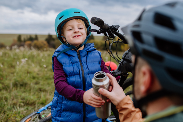 Portrait of father giving cup of warm tea to his son. Happy family resting, having snack during bike trail raiding in an autumn mountains.