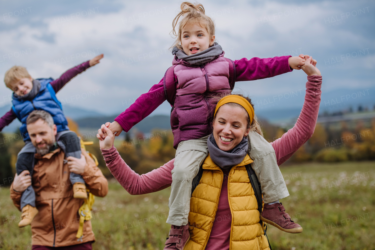 Happy parents with their little kids on piggyback at autumn walk, in the middle of colourfull nature. Concept of a healthy lifestyle. Carrying children on shoulders during the hike.