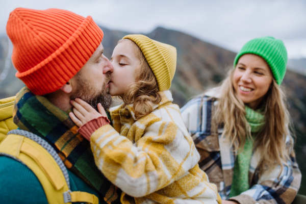 Portrait of happy family of mother, father and daughter on kid friendly hike in an autumn mountains. Little girl giving kiss to her father.