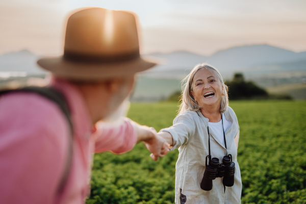 Senior couple standing in the middle of meadow and holding hands during beautiful sunset. Elderly woman with binoculars having romantic time with husband at vacation in mountains. Follow me photography.