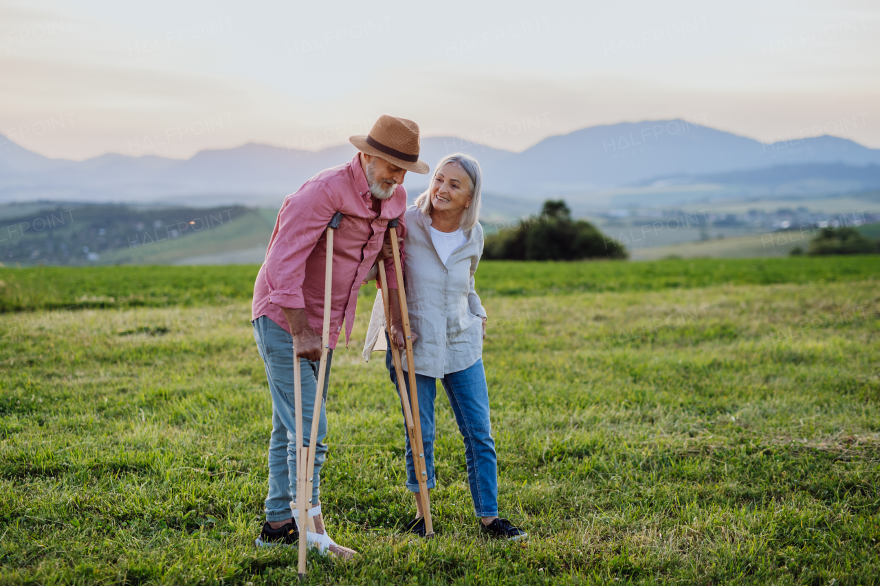 Senior man with broken leg and crutches standing in the the middle of meadow. Recovery, rehabilitation after injury or surgery in the nature. Wife helping husband with walking during easy walk on their vacation. Concept of traveling with physical disabilities.