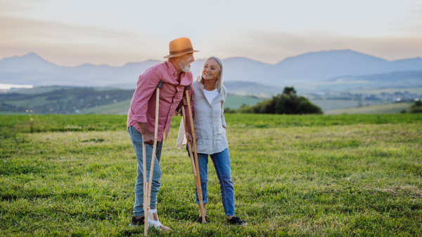 Senior man with broken leg and crutches standing in the the middle of meadow. Recovery, rehabilitation after injury or surgery in the nature. Wife helping husband with walking during easy walk on their vacation.