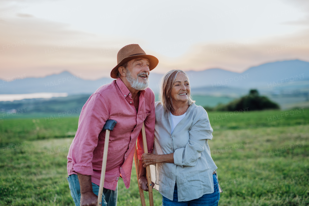 Senior man with broken leg and crutches standing in the the middle of meadow. Recovery, rehabilitation after injury or surgery in the nature. Wife helping husband with walking during easy walk on their vacation. Concept of traveling with physical disabilities.