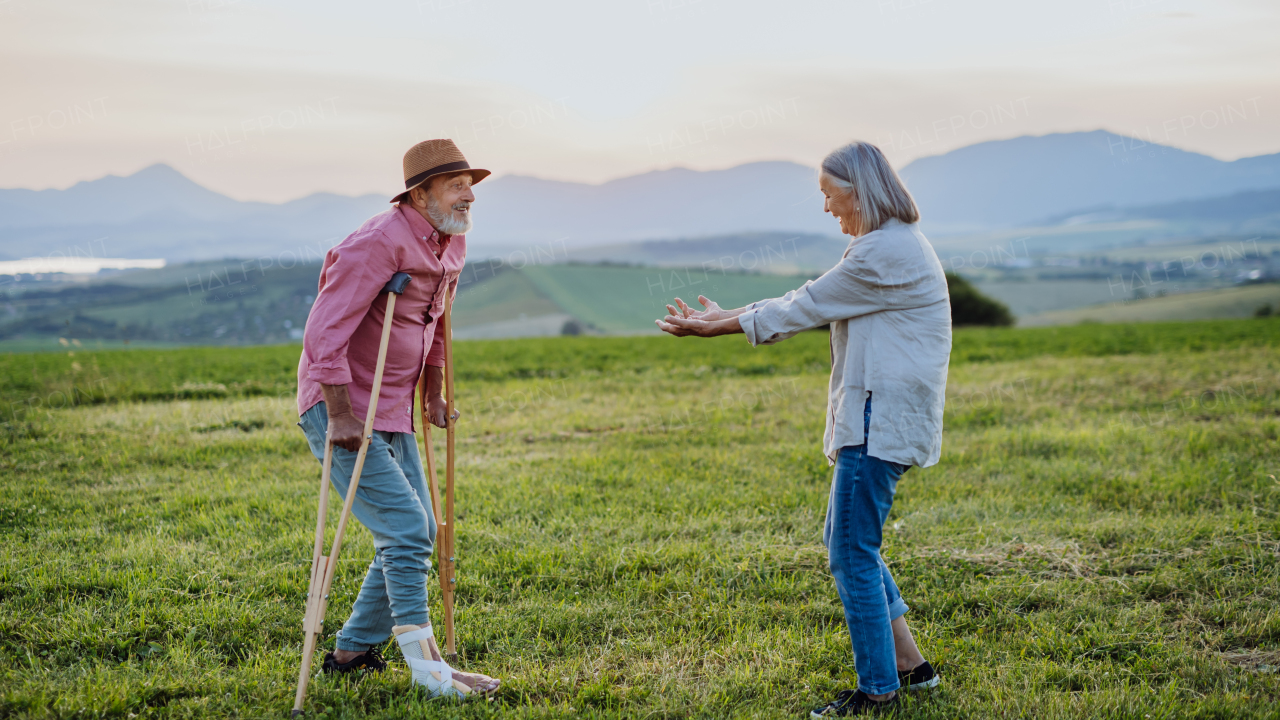 Senior man with broken leg and crutches standing in the the middle of meadow. Recovery, rehabilitation after injury or surgery in the nature. Wife helping husband with walking during easy walk on their vacation.