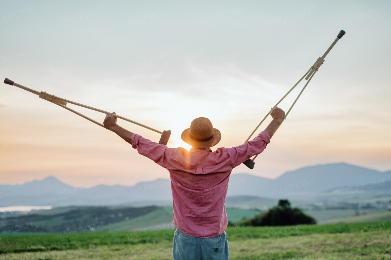 Rear view of senior man holding crutches in the air, he is happy abtou his recovery. Recovery, rehabilitation after injury or surgery in the nature. Concept of recovery and physical therapy