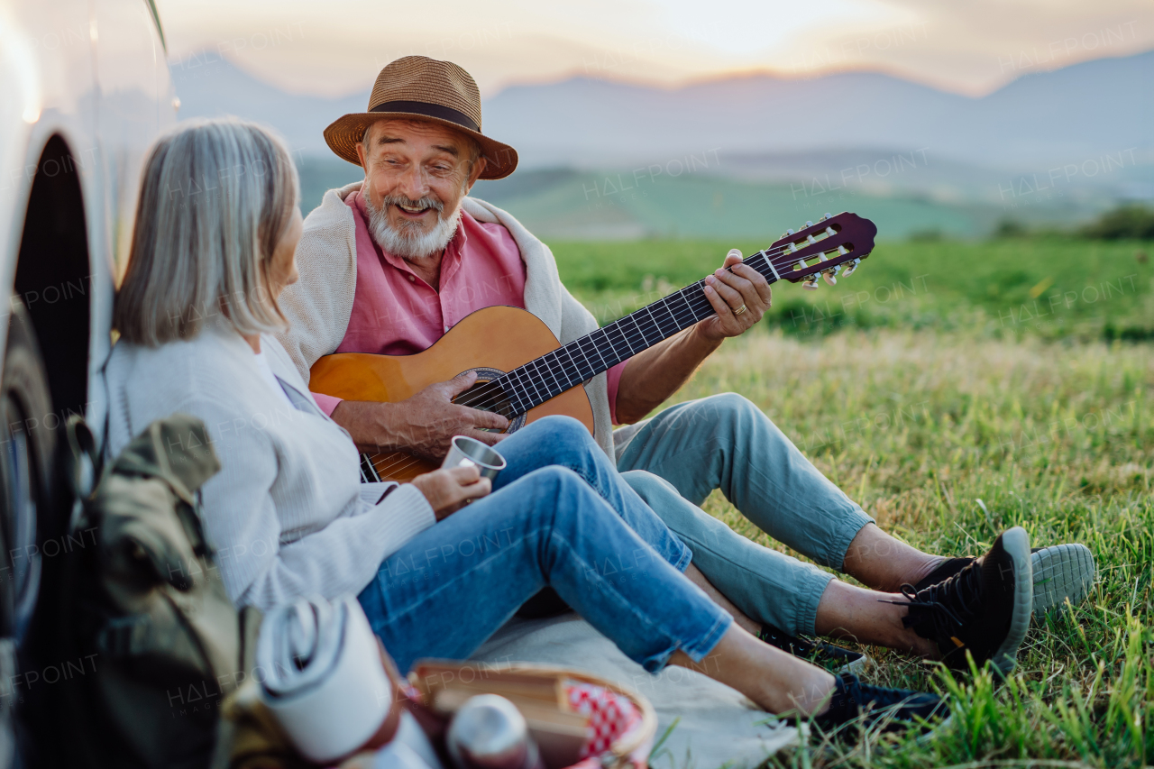 Potrait of senior couple sitting by ste car and playing guitar after long drive during their roadtrip. Elderly spouses at roadtrip enjoying serene landscape of High Tatras behind them.