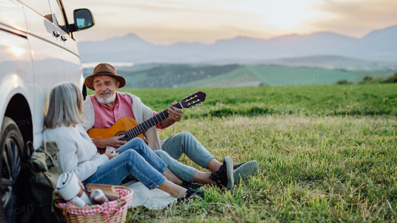 Potrait of husband playing the guitar, serenading his wife. Elderly spouses at autumn roadtrip watching the sun set behind High Tatras. Autumn banner with copy space.