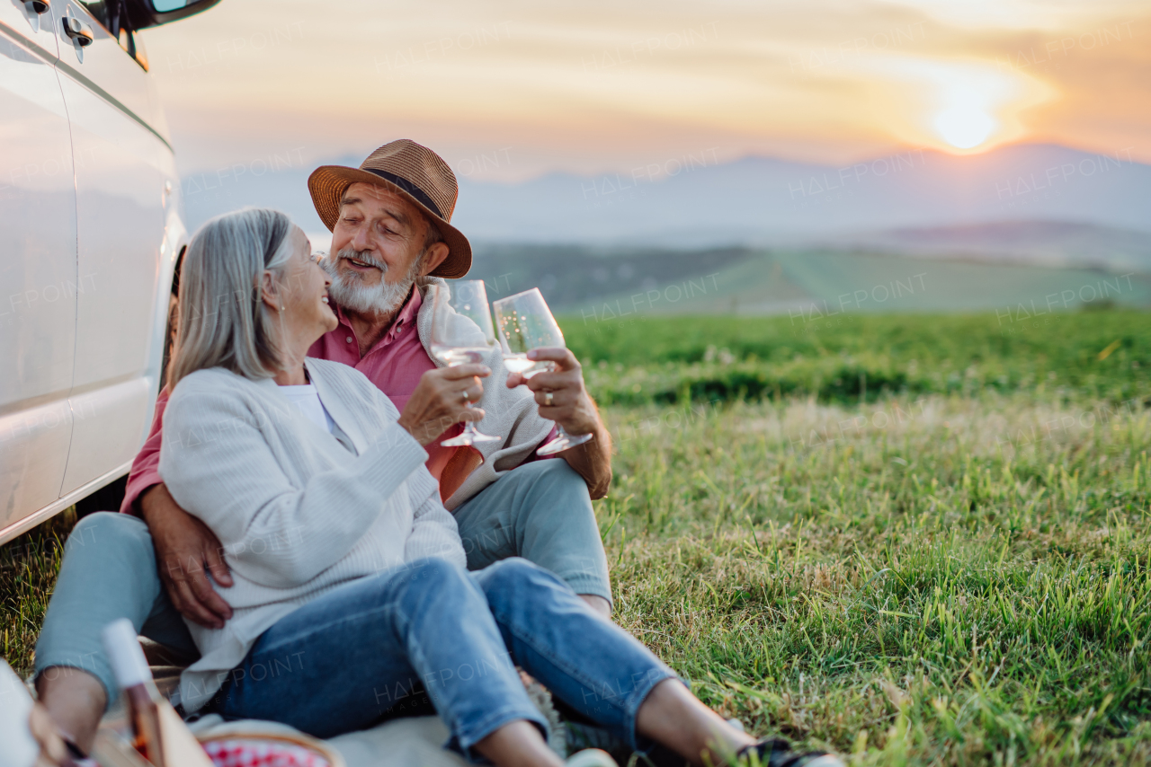 Potrait of senior couple sitting by ste car and drinking wine after long drive during their roadtrip. Elderly spouses at autumn roadtrip watching the sun set behind High Tatras.