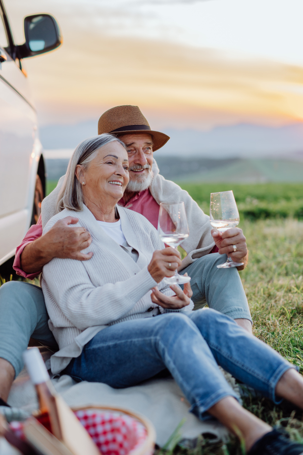 Potrait of senior couple sitting by ste car and drinking vine after long drive during their roadtrip. Elderly spouses at roadtrip enjoying serene landscape of High Tatras behind them.