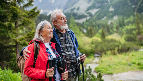 Active elderly couple hiking together in autumn mountains, on senior friendly trail. Husband and wife getting rest during hike, enjoying nature. Senior tourist with backpack using trekking poles for stability.