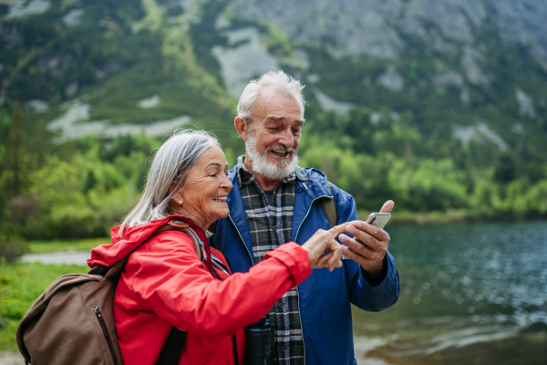 Active elderly couple hiking together in autumn mountains, on senior-friendly trail. Husband and wife looking at a map on a smartphone, searching for a hiking trail.