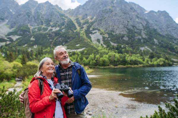 Potrait of active senior woman looking through binoculars at nature. Elderly couple hiking in autumn mountains on senior friendly trail. Senior tourist with backpack using trekking poles for stability.