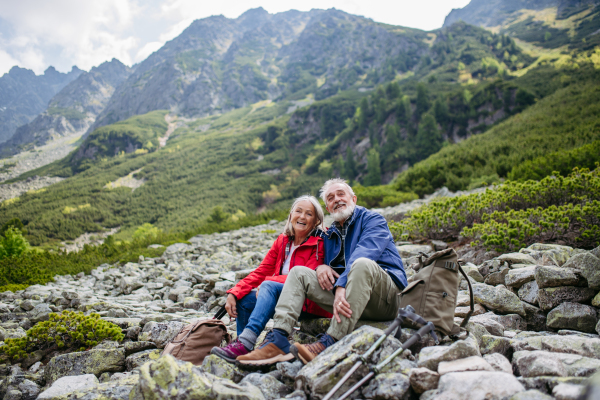 Active elderly couple hiking together in autumn mountains, on senior friendly trail. Husband and wife getting rest during hike, enjoying nature. Senior tourist with backpack using trekking poles for stability.