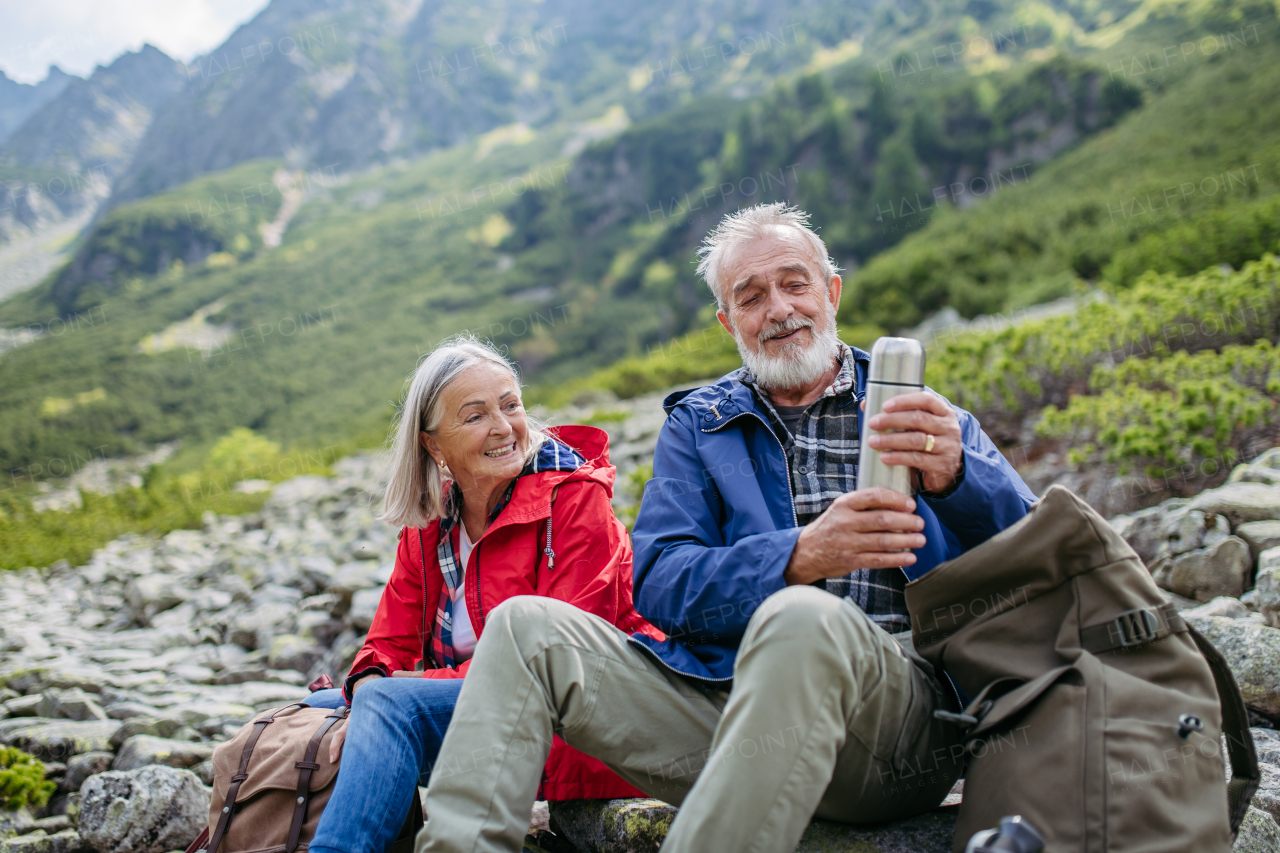 Active elderly couple hiking together in autumn mountains, on senior friendly trail. Husband and wife rehydrating, enjoying warm tea, coffee from thermos. Senior tourist with backpack getting rest during hike.