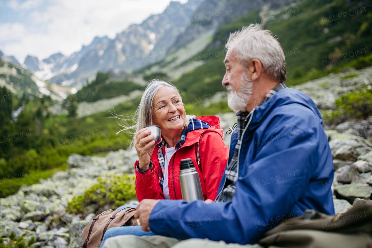 Active elderly couple hiking together in autumn mountains, on senior friendly trail. Husband and wife rehydrating, enjoying warm tea, coffee from thermos. Senior tourist with backpack getting rest during hike.