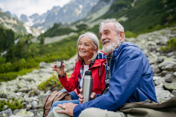 Active elderly couple hiking together in autumn mountains, on senior friendly trail. Husband and wife rehydrating, enjoying warm tea, coffee from thermos. Senior tourist with backpack getting rest during hike.