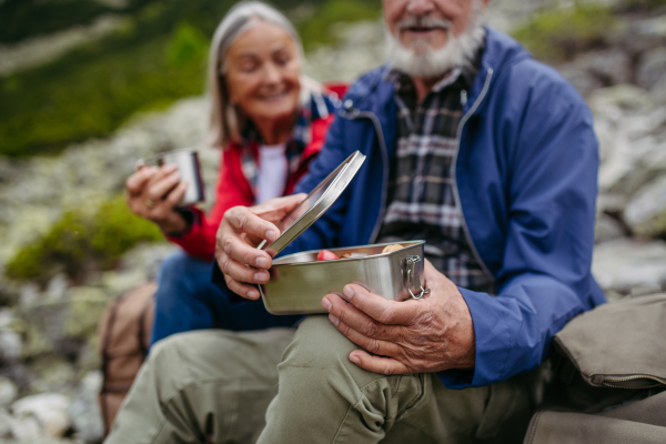 Active elderly couple hiking together in autumn mountains, on senior friendly trail. Husband and wife having healthy snack, to get energy. Senior tourist with backpacks resting during hike.