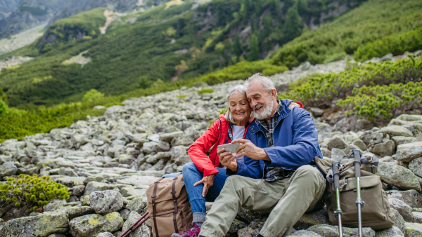 Active elderly couple hiking together in autumn mountains, on senior friendly trail. Husband and wife taking selfie with smartphone. Senior tourist with backpacks getting rest during hike.