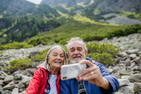 Active elderly couple hiking together in autumn mountains, on senior friendly trail. Husband and wife taking selfie with smartphone. Senior tourist with backpacks getting rest during hike.