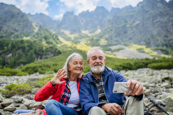 Active elderly couple hiking together in autumn mountains, on senior friendly trail. Husband and wife taking selfie with smartphone. Senior tourist with backpacks getting rest during hike.