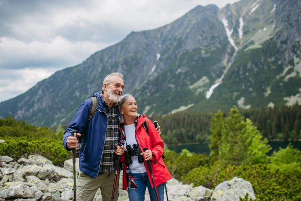 Waist up shot of active elderly couple hiking together in autumn mountains, on senior friendly trail. Husband and wife getting rest during hike, enjoying nature. Senior tourist with backpack using trekking poles for stability.