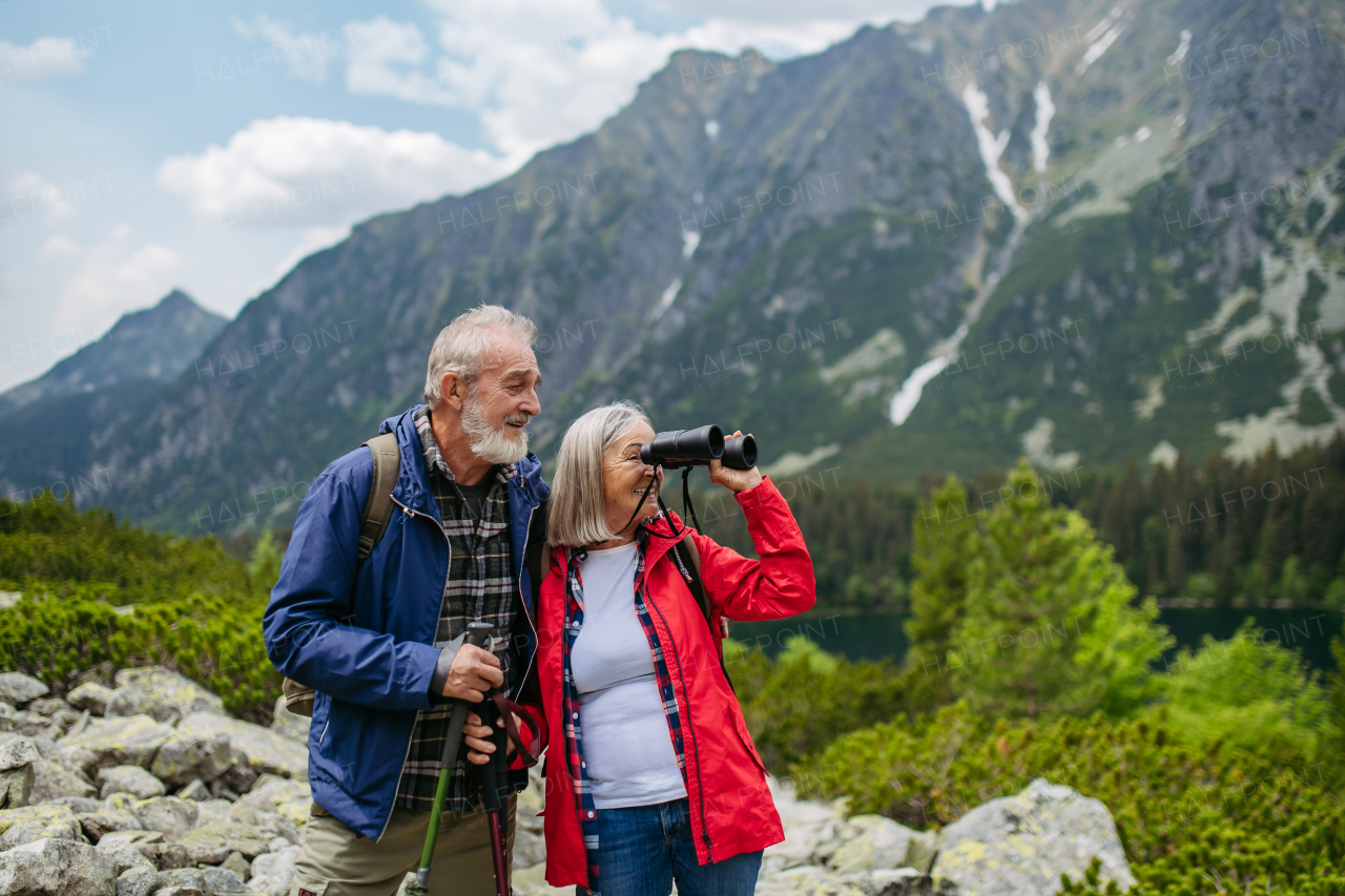 Potrait of active senior woman looking through binoculars at nature. Elderly couple hiking in autumn mountains. on senior friendly trail. Senior tourist with backpack using trekking poles for stability.