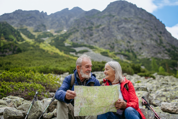 Active elderly couple hiking together in autumn mountains, on senior friendly trail. Senior spouses on the vacation in the mountains celebrating anniversary. Senior tourists sitting on rocks and looking at the route on a map.