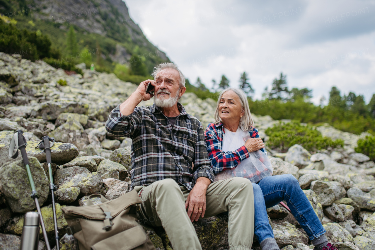 Senior tourists sitting on rocks and looking at the distant nature through binoculars. Active elderly couple hiking together in autumn mountains, on senior friendly trail. Senior spouses on the vacation in the mountains celebrating anniversary.