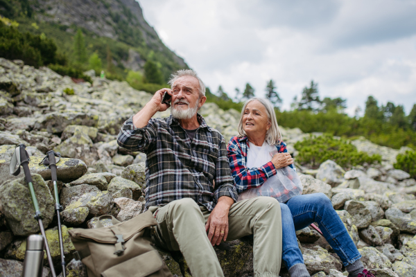 Senior tourists sitting on rocks and looking at the distant nature through binoculars. Active elderly couple hiking together in autumn mountains, on senior friendly trail. Senior spouses on the vacation in the mountains celebrating anniversary.