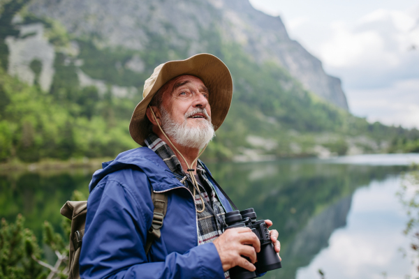 Potrait of active senior man hiking in autumn mountains, on senior friendly trail. Senior tourist with backpack looking through binoculars.