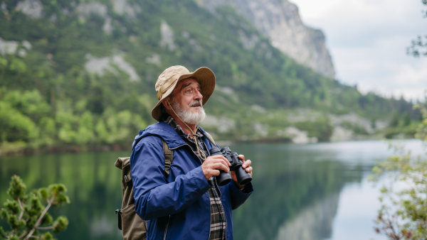 Potrait of active senior man hiking in autumn mountains, on senior friendly trail. Senior tourist with backpack looking through binoculars.