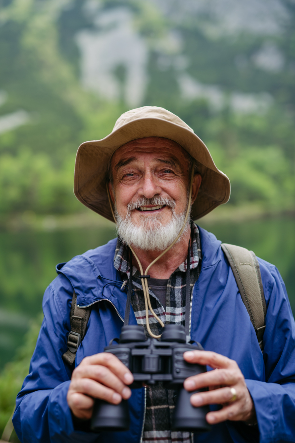 Potrait of active senior man hiking in autumn mountains, on senior friendly trail. Senior tourist with backpack looking through binoculars.