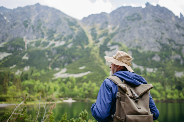 Rear view of active senior man hiking in autumn mountains, on senior friendly trail. Senior tourist with backpack looking through binoculars.