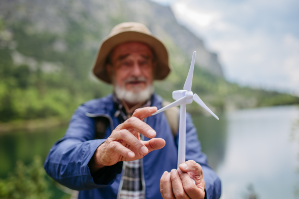 Potrait of active senior tourist holding a model of wind turbine. Senior man with backpack spinning the blade of turbine. Concept of renewable, green energy and sustainable lifestyle.